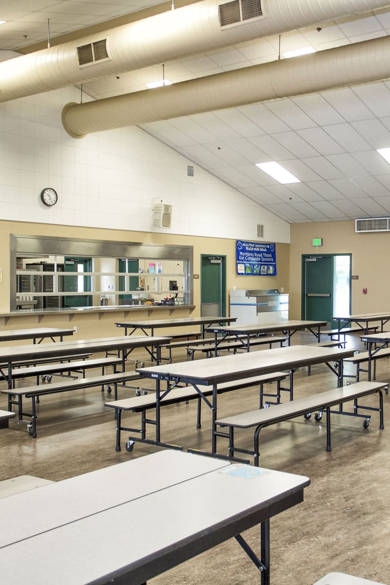School cafeteria with rows of tables and a wall-mounted display, the space Marshall Industries equips with sound masking and digital menu boards.